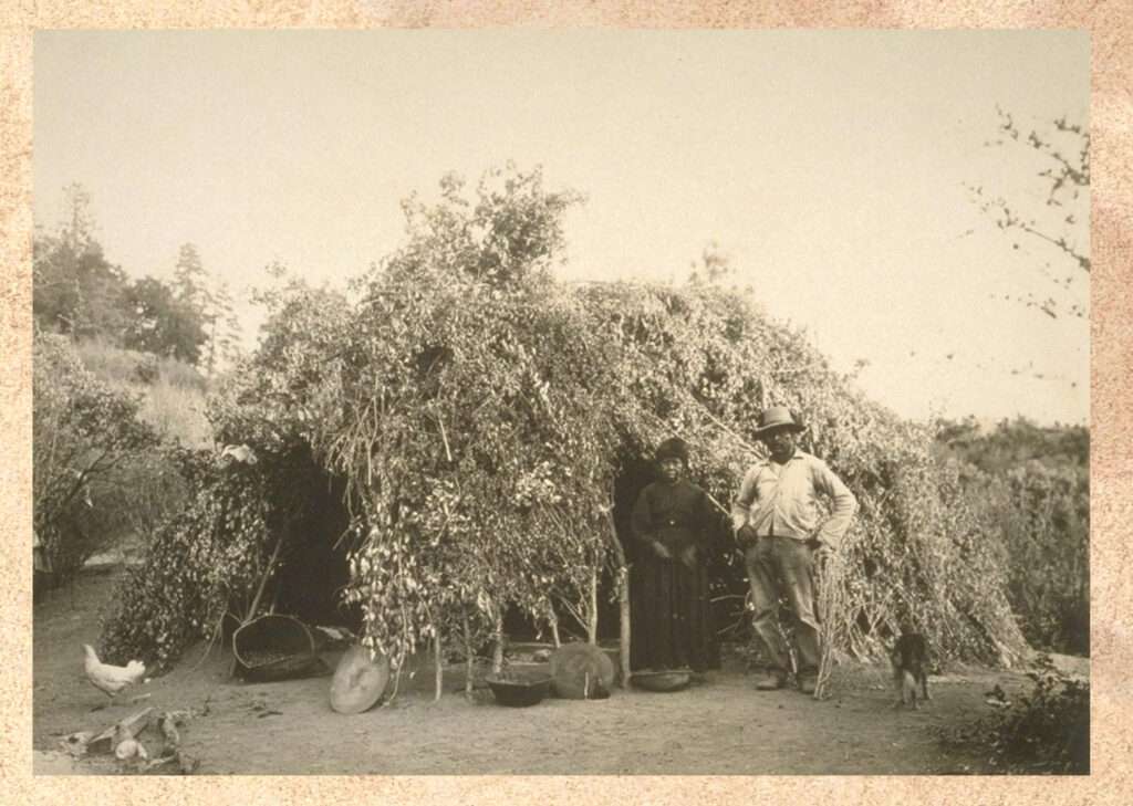 old photo of a hut made out of thatch and sticks with man